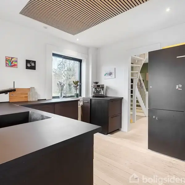 Et moderne køkken med mørke skabe og indbyggede apparater. Sort vask og vandhane på en køkkenø. Billedkunst på væggen. Vindue med udsigt til have. Trappe i baggrunden.