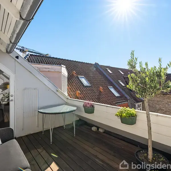 En træterrasse med en sort sofa og et rundt glasbord viser udsigten til røde hustage under en klar blå himmel. Planter i potter står langs rækværket.