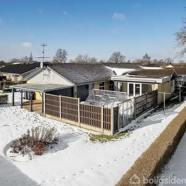 Et parcelhus med snedækket have står stille omgivet af hegn og stier i et villakvarter. Et drivhus ses ved siden af huset under en klar blå himmel.