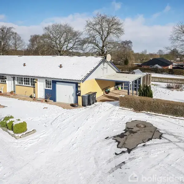 Et rækkehus med hvidt og gult ydre står i et snedækket område med bar træer og klare himmel. Grunden er omkranset af en hæk, og der er sneklædte stier.