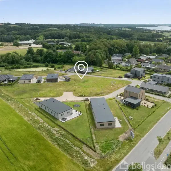 Et parcelhuskvarter viser spredte moderne huse placeret langs veje, med en markør, der angiver en specifik ejendom. Grønt landskab omgiver kvarteret, med skovområder og marker i baggrunden.