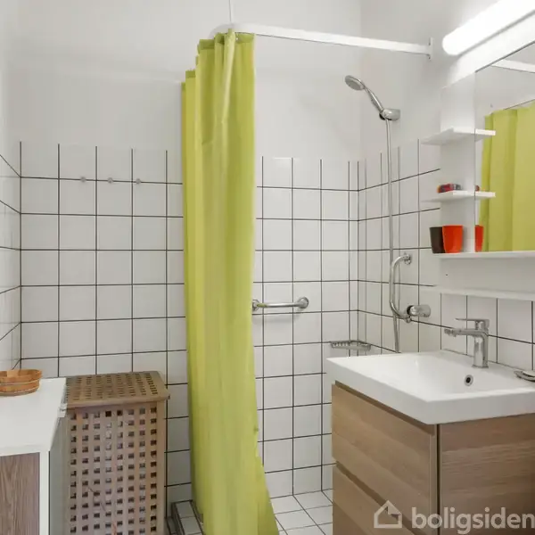 Brusekabine med hvidt flisebelagt væg og limegrønt badeforhæng. Badeværelsesskab med vask og spejl over samt hylde med to røde glas til højre. Træskab til venstre.