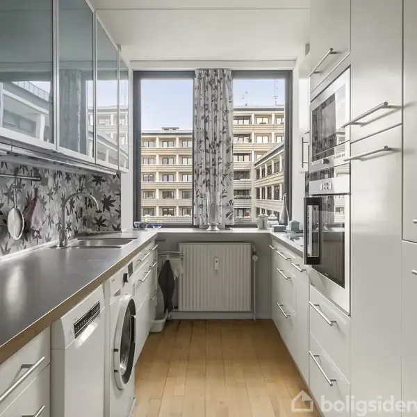 Et moderne køkken med hvidt interiør har skabe og vask, set mod et stort vindue, der afslører en udsigt til nabobygningen. Gardiner hænger delvist til siden.