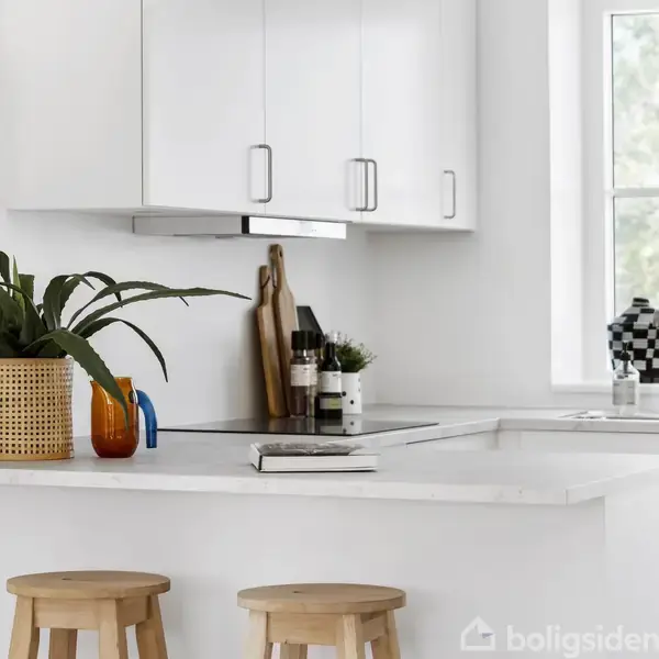 To træskamler står ved en hvid køkkenø med planter og køkkenredskaber. Bagved ses hvide skabe, en vindueskarm med en vase og et vindue der giver naturligt lys.