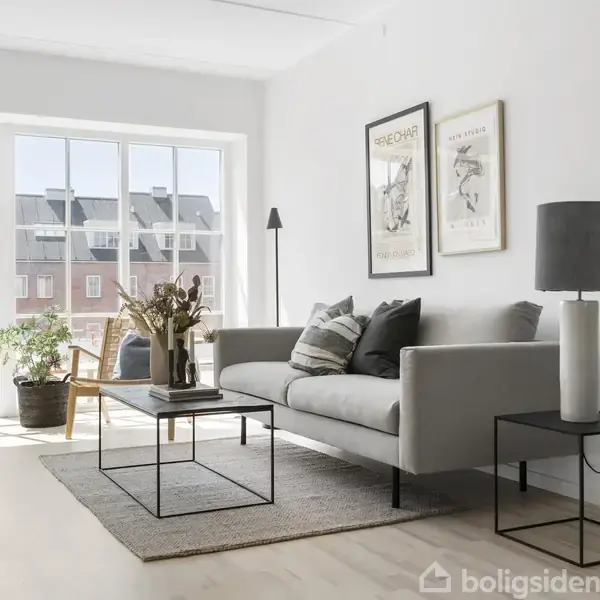 Stue med grå sofa står ved et stort vindue; to billeder hænger over sofaen. Et sofabord og lampe er ved siden af. Udsigt til anden bygning udenfor.