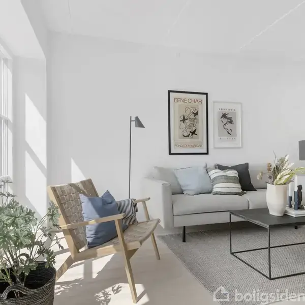 Sofa med puder står i stue, omgivet af en lænestol og et sofabord med vase og bøger. Store vinduer bringer lys ind. Plakater på væggen viser kunstmotiver.