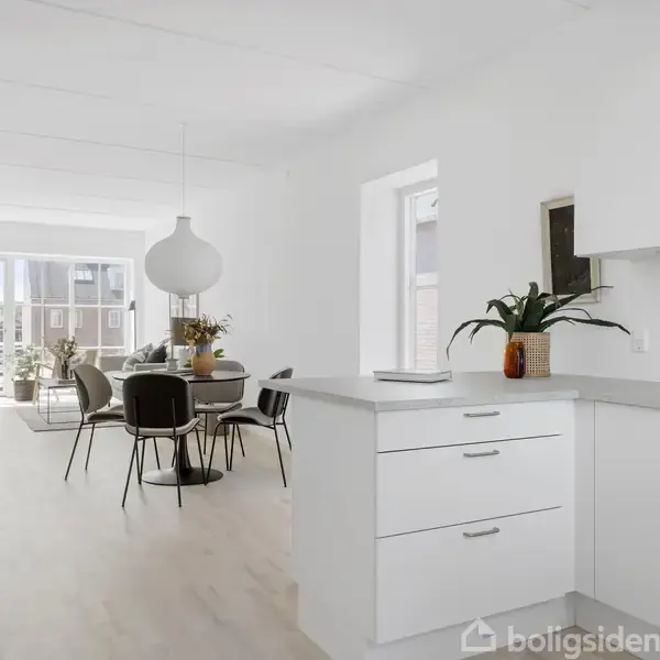 Et moderne, lyst køkken med hvide skabe, hvor en plante står på bordpladen. I baggrunden er der et spisebord med stole, og store vinduer slipper masser af lys ind.