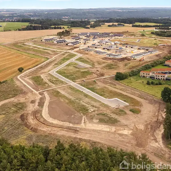En byggeplads med udlagte veje under udvikling omkranset af marker og huse i det fjerne, muligvis en kommende boligområde i et landligt landskab.