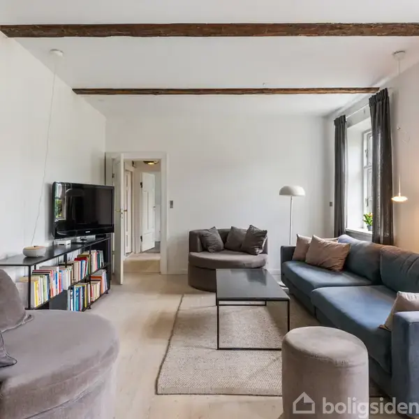 Moderne stue med to sofaer og en lænestol, placeret omkring et sofabord. Væggene er hvide med et farverigt maleri. Rummet har store vinduer og en bogreol ved TV'et.