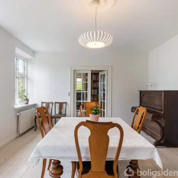 Et spisebord med hvid dug er centralt placeret omgivet af klassiske træstole; bagved ses et mørkt klaver, omgivet af lyse vægge og store vinduer med udsigt til grønne omgivelser.