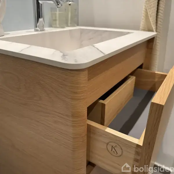 Trævaskeskab med en åben skuffe under en flade af hvid marmor. I baggrunden ses væggen og hjørnet af et håndklæde.