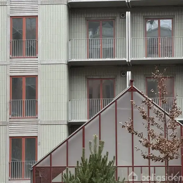 Bygning med flere etager og altaner, malet i lyse farver med rødmalede vinduesrammer. En trekantet overdækning med træer står foran, hvilket giver en moderne, urban atmosfære.