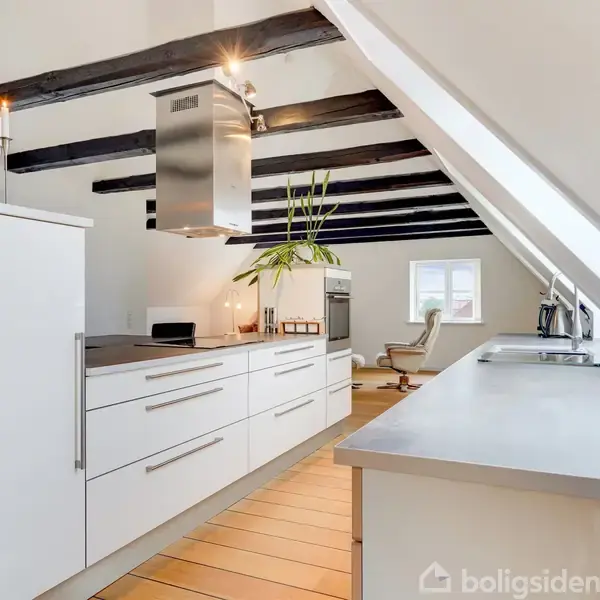 Et moderne køkken med hvide skabe og integreret ovn, placeret under et loft med synlige bjælker. Et plante står på bordpladen ved siden af et tændt stearinlys.