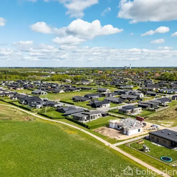 Boligområde ses fra luften med moderne, ensartede huse arrangeret i rækker, omkranset af grønne områder og en klar blå himmel med få skyer.