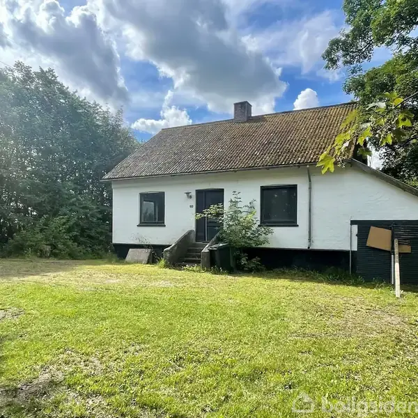 Et hvidt hus står i en solbeskinnet have omgivet af træer. Huset har en skråtag og en lille trappe op til hoveddøren. Skyer er synlige på himlen.