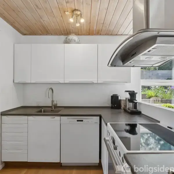 Køkken med hvide skabe og stålvask. Elkedel og kaffemaskine står på bordet. Kogeplade med emhætte tæt på et vindue. Træloft og døråbning til et andet rum.