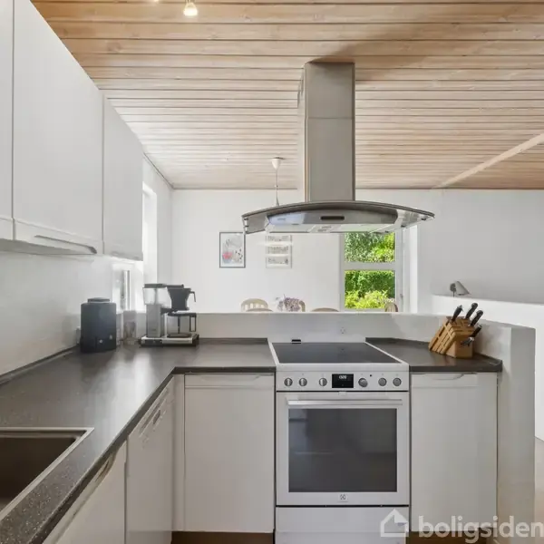 Et moderne køkken med hvidkalkede skabe og grå bordplade. En indbygget ovn og komfur står centralt under en emhætte. I baggrunden ses et vindue med udsigt til grønne træer.