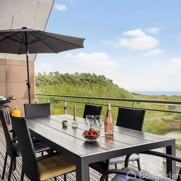 Et udendørs bord med sorte stole står på en terrasse med udsigt over grønne klitter og hav. På bordet er der glas, en flaske, og frugter, samt en paraply i nærheden.
