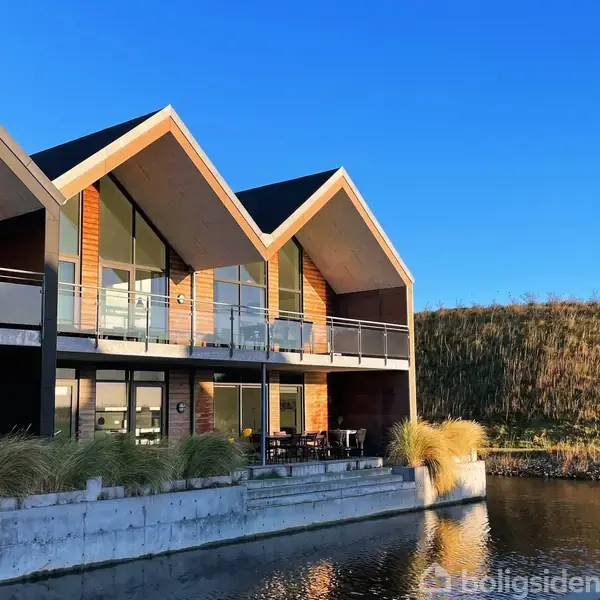 En moderne bolig med trekantede tage og store vinduespartier reflekteres i det stille vand foran. Den står i et åbent, grønt miljø med en blå himmel i baggrunden.
