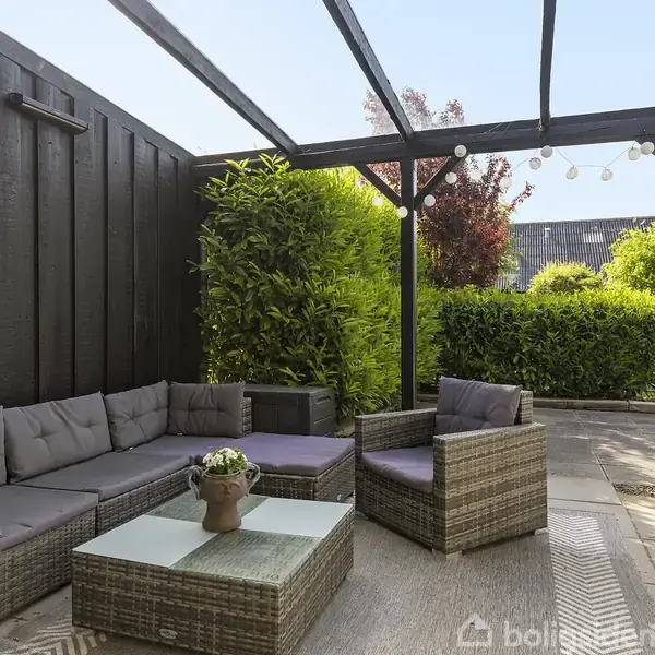Terrassemøbler i flettet design står under en overdækket terrasse. Hynderne er grå, og et sofabord med en blomsterdekoration er placeret i midten. Omkranset af grønne buske.