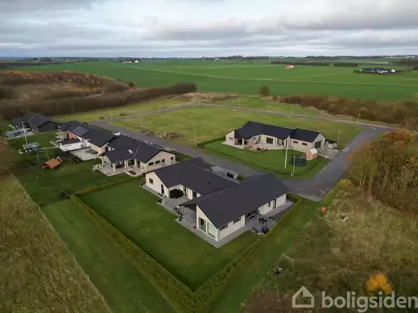 Huse med sorte tage og velplejede græsplæner ligger i et rækkehusområde omgivet af grønne marker og åbne vidder under en overskyet himmel.