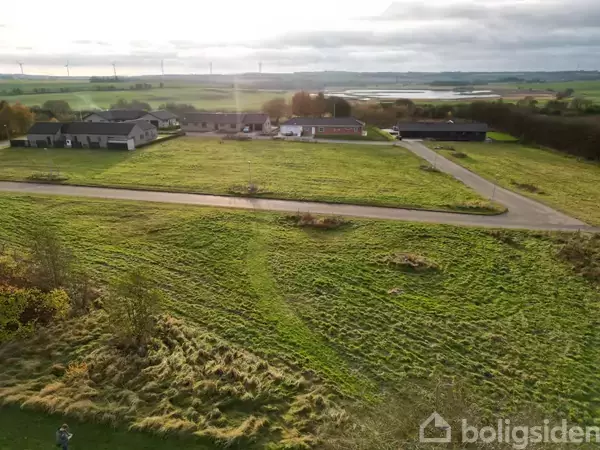 Grøn mark strækker sig foran et par enfamiliehuse ved en vej. Vindmøller ses i horisonten under en lys, overskyet himmel.