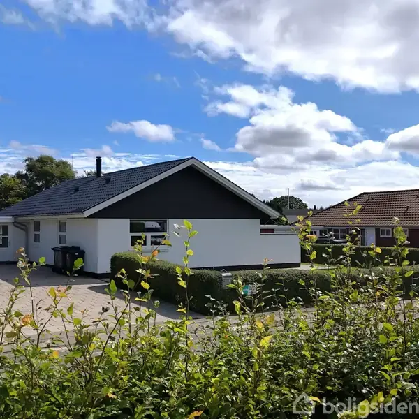 Et hvidt hus med sort tag står stille i et beboelseskvarter, omgivet af velplejede hække og en flisebelagt indkørsel. Blå himmel og spredte skyer ses i baggrunden.