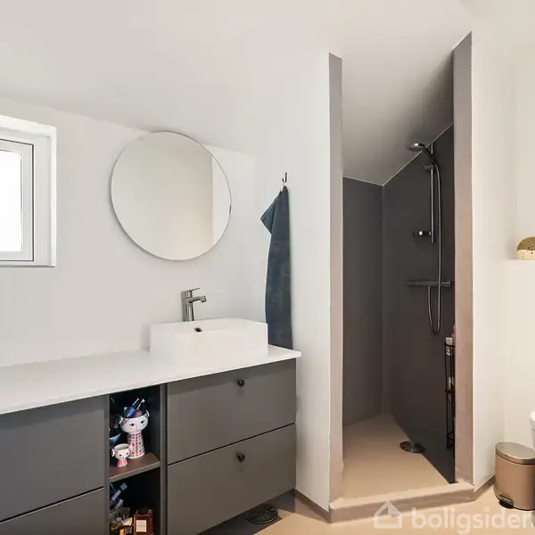 Badeværelse med hvid vask på grå skab, rund spejl over. Bruser til højre ved siden af en væghængt toiletkumme. Et lille vindue og plante er i baggrunden.