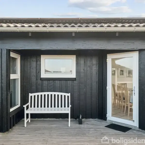 Hvid bænk står stille foran sort træhus med store vinduer og en glasdør i sollys. Indenfor ses et spisebord og stole i lyse omgivelser.