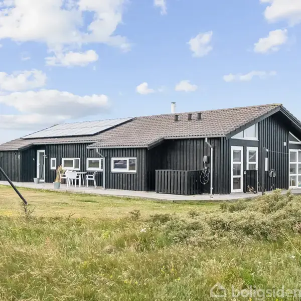 Et sort træhus med hvide vinduer står på en græsplæne. En gynge er til venstre. Skyer ses mod en blå himmel.