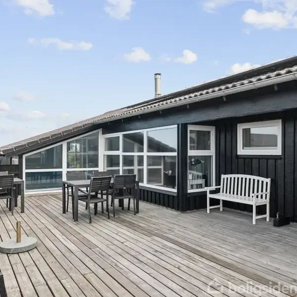 En stor terrasse med træbrædder har sorte møbler, herunder borde og stole. Den er forbundet til et moderne sortmalet hus med store vinduer, under en blå himmel med lette skyer.