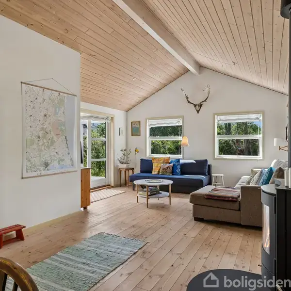 En stue med en brændeovn, to sofaer ved vinduer, et hjørnekabinet, kort på væggen og et rummeligt træloft. Udsigt til skov gennem vinduer.