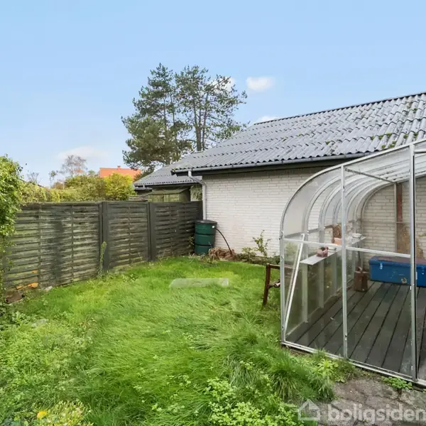 Et lille drivhus af glas står på en træterrasse ved siden af et hvidt murstenshus. Omkring drivhuset er en grøn have med græs og træer, og en træhegn markerer grænsen.
