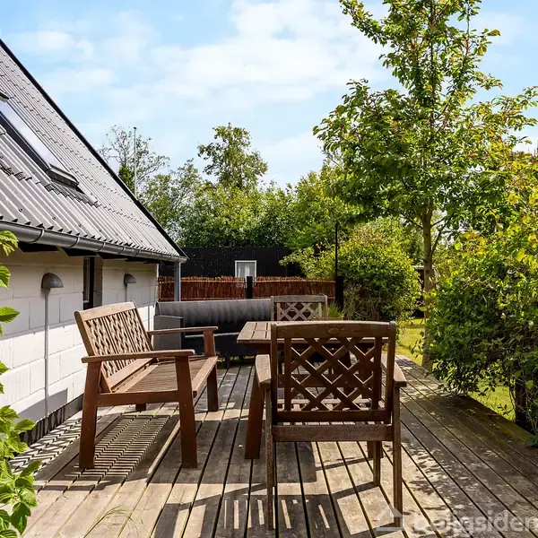 Terrasse med træbænke og træbord står på et trædæk. De omgives af frodigt grønne planter og et hvidt hus med et tag med ovenlysvinduer. Solrigt vejr og blå himmel.