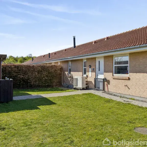 Rækkehus med rødt tag ligger i en velholdt have med græsplæne og flisebelægning. Ved siden af huset står en træbygning. Himmel og hæk udgør baggrunden.
