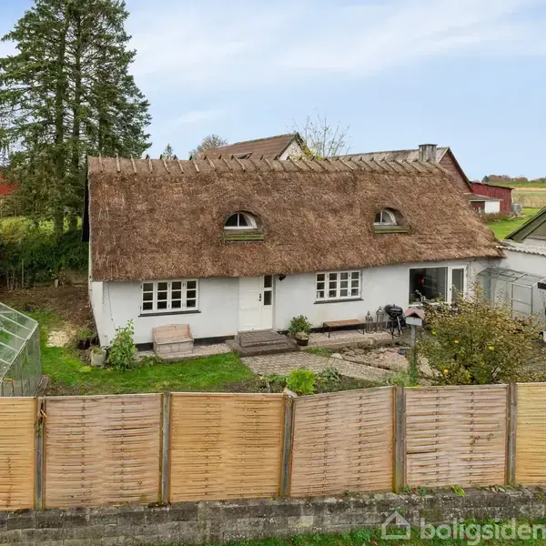Et hvidt hus med stråtag står blandt en have med stakit og drivhus. Træer og bygninger omkranser det, mens det åbne landskab ses bagved.