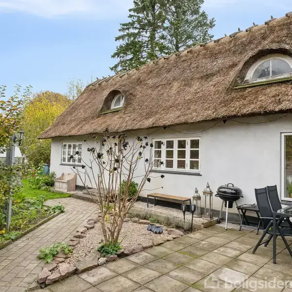 Stråtækt hus med to runde vinduer på taget. Haven er anlagt med fliser og havemøbler. Træer og buske omgiver huset, der fremstår idyllisk og naturskønt.