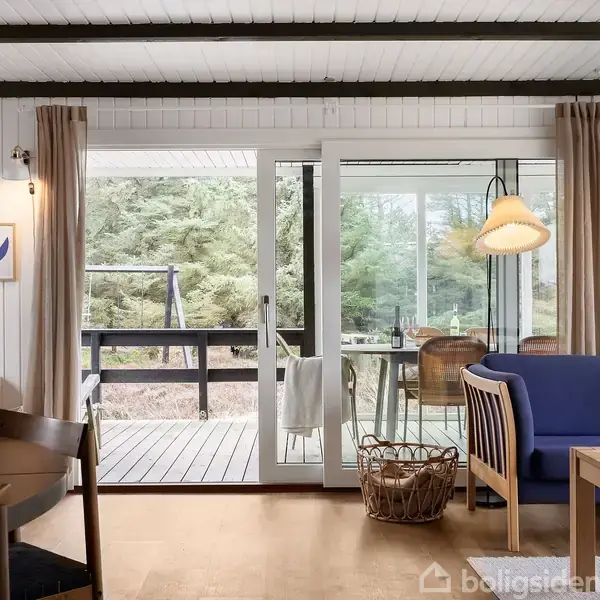 Stue med træmøbler, hvor en sofa og rummeligt bord er centralt placeret. Store glasdøre åbner til en terrasse med udsigt til skovområde. Vægpynt viser ord "Gin Gin."