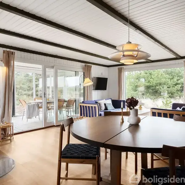 Stue med rund træspisebord, omgivet af stole, pejs i hjørnet med ild, store vinduer ud til terrasse. Blå og hvide sofaer, kunst på væggene og grøn udsigt udenfor.