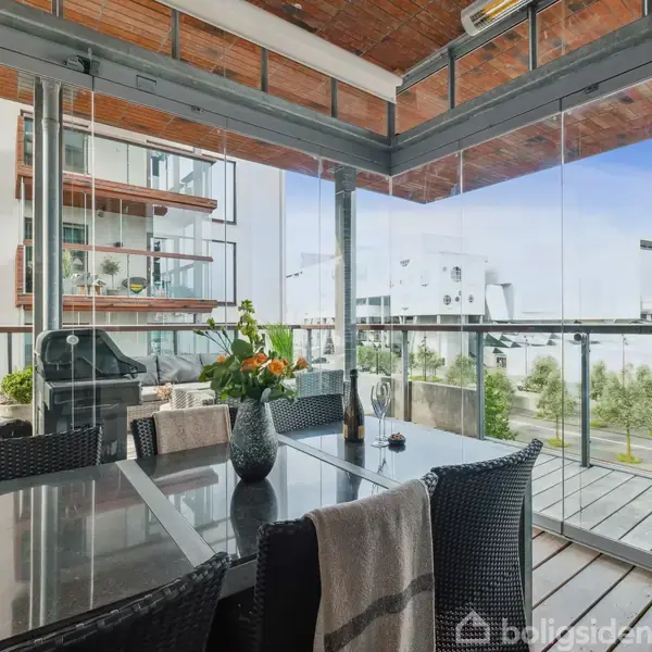 Et bord med sorte fletstole står på en glasterrasse med udsigt over moderne bygninger. Der er blomster på bordet, og omgivelserne er hyggelige og lyse.