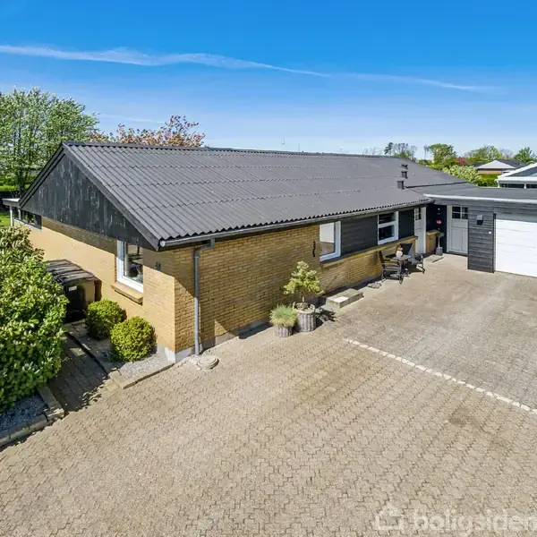 Et gult murstenshus med et sort tag ligger ved siden af en garage i en brolagt indkørsel omgivet af buske og træer under en klar blå himmel.