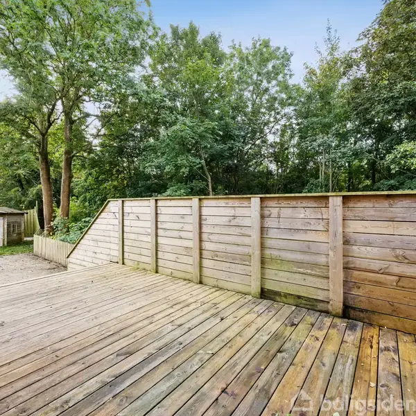 En træterrasse strækker sig ud fra et hus, omgivet af en lav træhegn. I baggrunden ses en frodig have med græs, træer og en træhytte. Ingen tekst fundet.