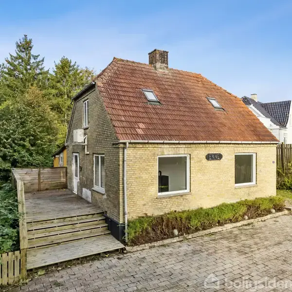 Et murstenshus med rødt tegltag står stille i en skovklædt have. Foran huset er en flisebelagt gårdsplads og til højre en træterrasse omgivet af træer.