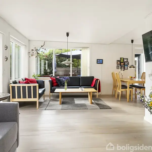 Stue med moderne møblering; sorte sofaer og træbord står på trægulv. En fladskærm hænger på væggen. Store vinduer giver udsigt til en terrasse. Blomster står i hjørnet.