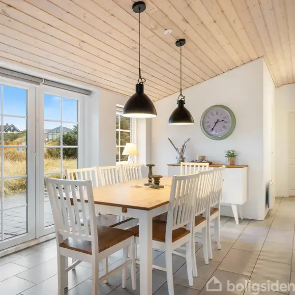 Spisebord med seks hvide stole står i en lys spisestue. To sorte lamper hænger over bordet. Store vinduer viser en terrasse og natur. En vægur findes på væggen.