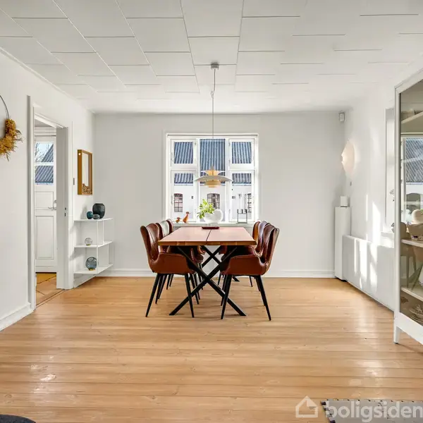 Spisebord med seks brune stole står i et lyst rum med trægulv. Bordet er belyst af en hængelampe. En glasskab står til højre, og et vindue er i baggrunden.