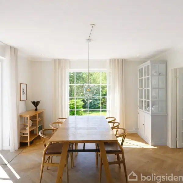 Spisebord, omkranset af stole, står centralt i det lyse spisestue. Sollys stråler ind fra en stor vindue, omgivet af lyse gardiner, med udsigt til grønne omgivelser udenfor.