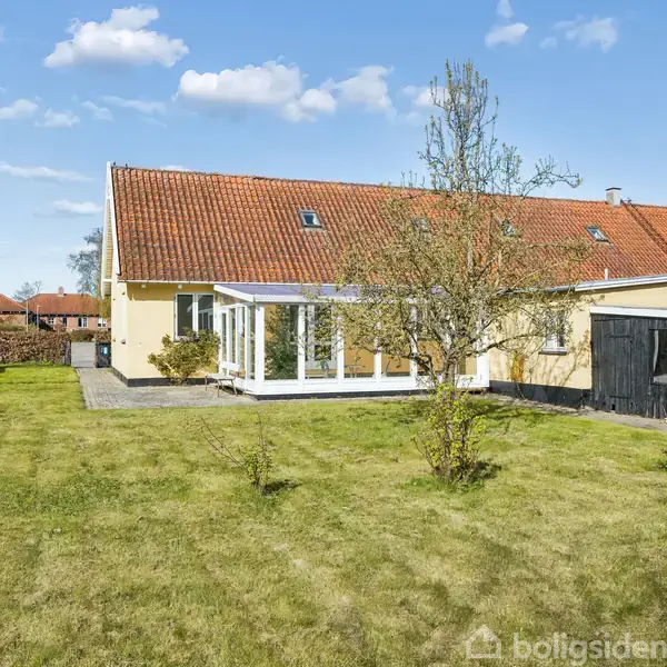 En gul villa med rød tagstenstag står i en solbeskinnet have. Huset har en udestue med glasfacade og en sort skur, omgivet af græsplæne og få træer.