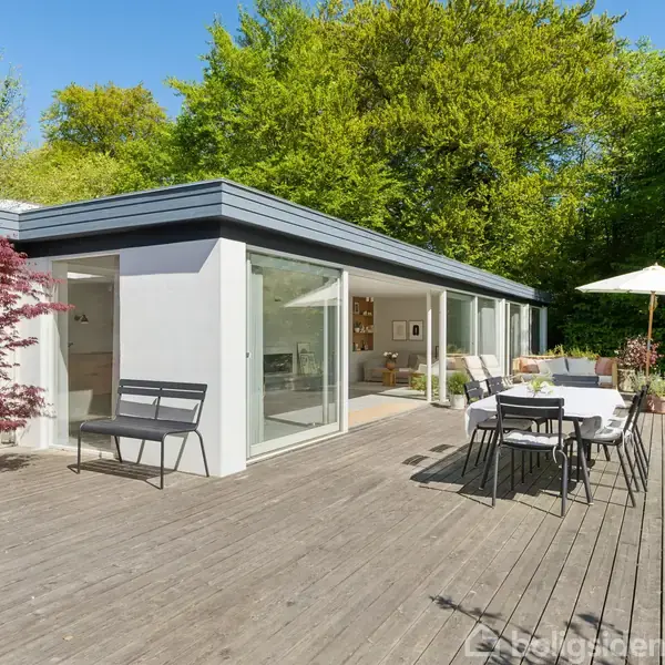 Et moderne hus med store glasdøre står i solen. En træterrasse med borde, stole og en parasol skaber en afslappende atmosfære omgivet af grønne træer og buske.