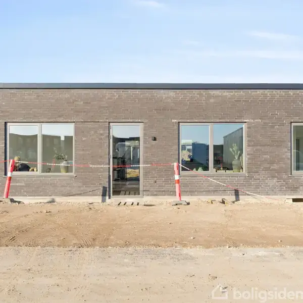 Moderne etplanshus af mørk mursten står stille på en byggeplads. Omgivet af jord og afspærringsbånd i et åbent, solrigt område. Tre store vinduer og en glasdør dominerer facaden.